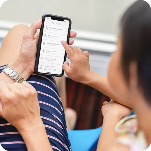 Person holding a smartphone displaying a "My Lessons" app interface on the Big Blue swim school app, with a finger touching a lesson entry, and another person partially visible to the right.