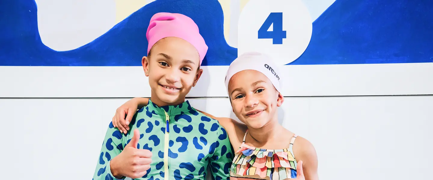 two young girls standing side by side in their swimsuits and swim caps smiling at the camera and giving a thumbs up