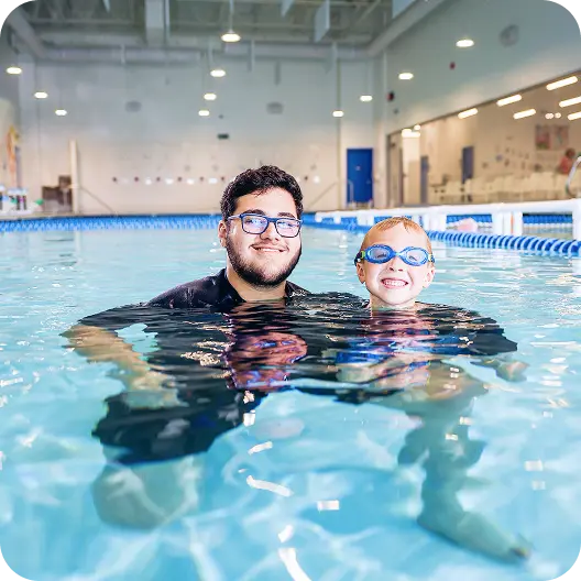 A young man with glasses and a small boy in goggles in a swimming pool, smiling and looking at the camera. They are both submerged up to their chests in the water.