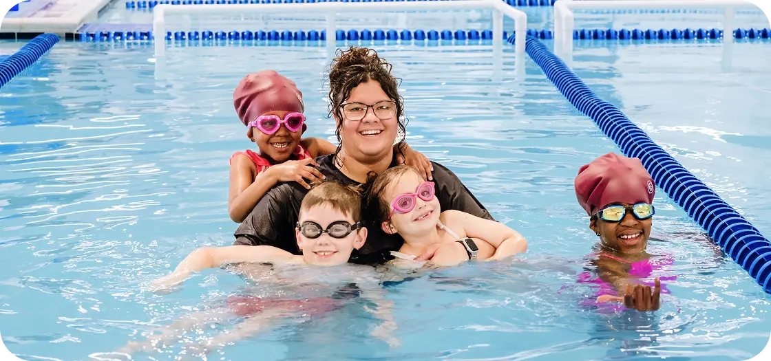 A swimming instructor and four children in a pool, all wearing goggles and swim caps, smiling at the camera.