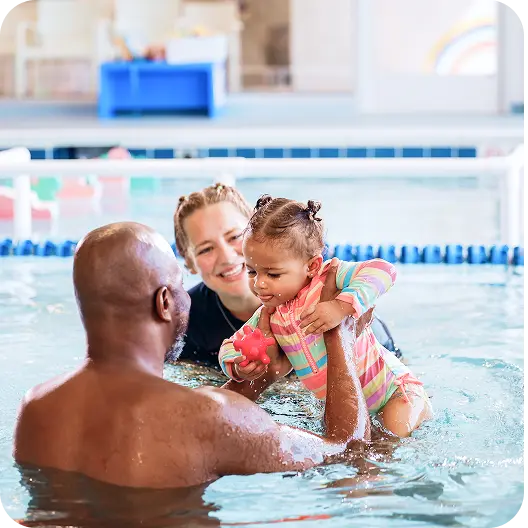 A man holds a smiling baby in a colorful striped swimsuit in a swimming pool, while a smiling swim instructor helps behind them