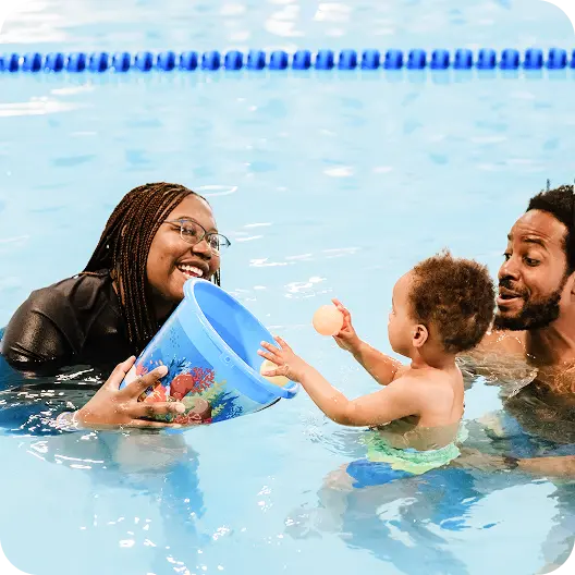 A smiling swimming instructor, a man, and a baby are in a pool. The instructor holds a blue bucket with sea life designs in the water, from which the baby reaches for a small, orange ball.