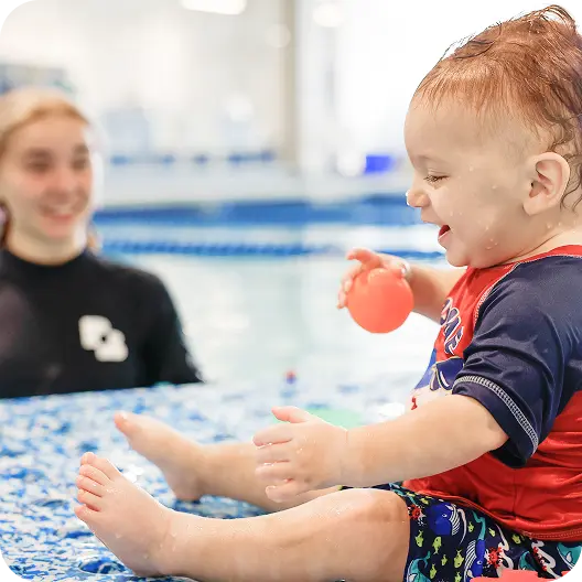A toddler in a red and blue rash guard and swim trunks laughs while sitting on a patterned mat near a swimming pool, holding an orange ball with a blurry instructor visible in the background.