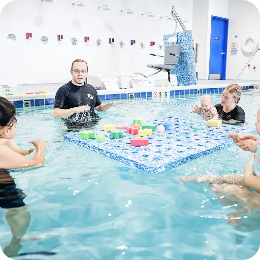 A swim instructor in a black shirt gestures while teaching a water class to babies and their parents in a swimming pool, with a large blue and white mat holding floating toys in the center.