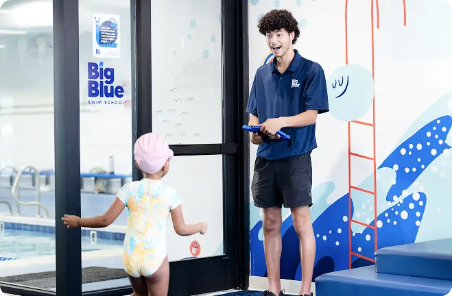 A Big Blue staff member is holding a tablet and smiling wide in the waiting room while a young girl runs to him in her swimsuit, swim cap, and goggles