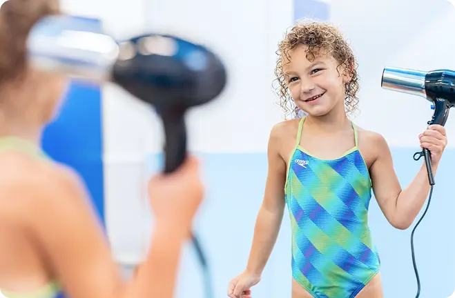 a young girl with curly hair and a blue and green swimsuit is using a hair dryer to dry her hair after a swim lesson
