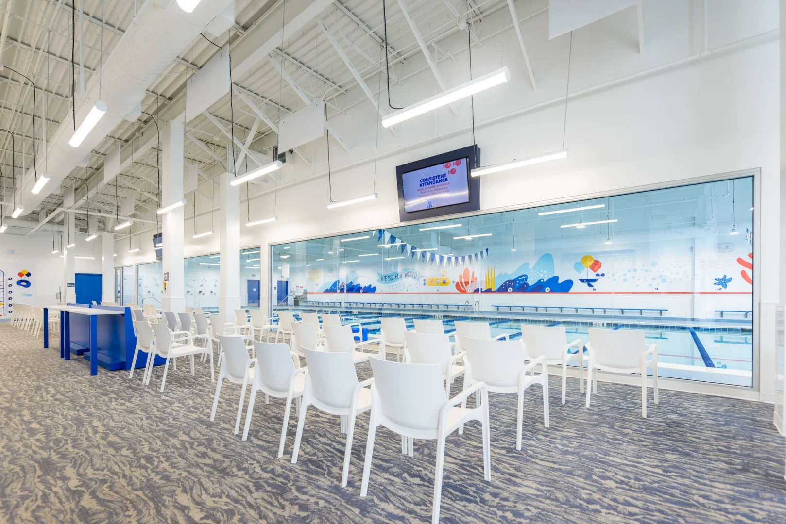 Interior view of Big Blue Swim School's waiting area, facing a swimming pool through a large window, featuring rows of white chairs arranged in front of the pool, a digital screen, and overhead lighting.