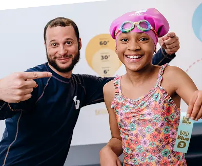 Smiling girl in a floral swimsuit and pink swim cap with goggles, holding a "Bold Blue 3" achievement ribbon, poses next to a smiling instructor wearing a dark blue shirt